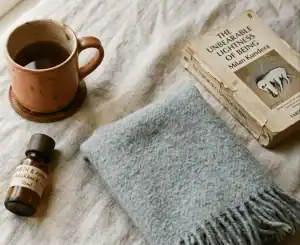 A mug, perfume oil bottle, a blanket, and a well-loved book laying on a tablecloth.